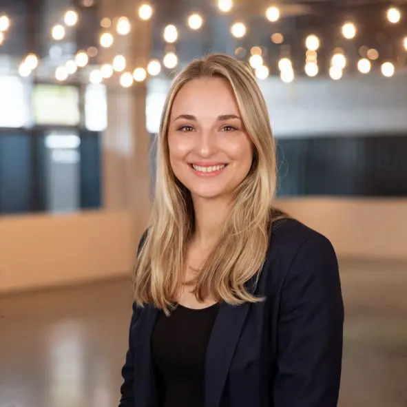 Eine junge Frau mit langen blonden Haaren, die einen dunklen Blazer und ein schwarzes Oberteil trägt, lächelt in die Kamera. Sie steht in einem modernen, gut beleuchteten Raum mit Lichterketten an der Decke und großen Fenstern im Hintergrund.