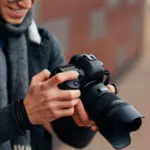 Eine Person, die eine digitale Spiegelreflexkamera in der Hand hält, Einstellungen vornimmt und dabei lächelt. Der Hintergrund ist unscharf, und die Person trägt einen Schal, eine Jacke und eine Brille.