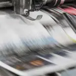 Nahaufnahme einer Druckerpresse in Aktion, mit unscharfen Papierbögen, die sich schnell unter einer Metallwalze bewegen und bunte Druckerzeugnisse produzieren.