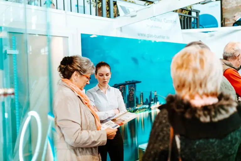 Eine Frau in einem weißen Hemd spricht mit einer älteren Frau, die an einem Messestand Broschüren in der Hand hält, während andere Personen in der Nähe stehen. Im Hintergrund ist ein großes Foto einer Stadtlandschaft zu sehen.