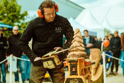 Ein Mann, der einen Gehörschutz und eine Schutzbrille trägt, schnitzt mit einer Kettensäge eine Baumskulptur aus Holz, wobei Holzspäne fliegen. Im Hintergrund sehen Menschen bei einer Veranstaltung im Freien zu, die in einer aktuellen Pressemitteilung vorgestellt wurde.