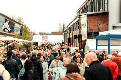 Eine große Menschenmenge schlendert durch einen Markt oder ein Festival im Freien, umgeben von Backsteingebäuden und Verkaufsständen unter bewölktem Himmel. Inmitten des geschäftigen Treibens sind bunte Fahnen und Regenschirme zu sehen, die eine lebhafte Atmosphäre schaffen, die einer Pressemitteilung würdig ist.