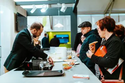 Ein Mann im Anzug demonstriert einer Gruppe von Personen, von denen einige Lebensmittel und Notizbücher in der Hand halten, das Kochen an einem Küchentisch in einem modernen Innenraum - perfekt für eine Pressemitteilung, die kulinarische Innovationen hervorhebt.