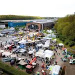 Luftaufnahme einer Freiluftveranstaltung mit vielen Zelten, Ständen und Fahrzeugen. Die Menschen flanieren zwischen den Auslagen auf einem großen Parkplatz in der Nähe von Industriegebäuden und Grünanlagen, im Hintergrund sind ein MediaMarkt und "Pressemitteilung"-Banner zu sehen.