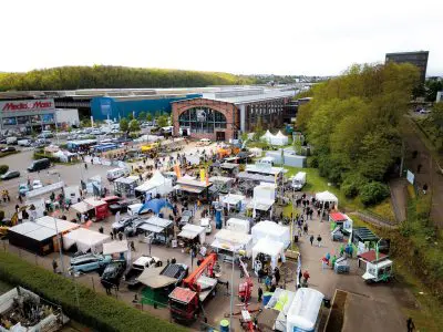 Luftaufnahme einer Freiluftveranstaltung mit vielen Zelten, Ständen und Fahrzeugen. Die Menschen flanieren zwischen den Auslagen auf einem großen Parkplatz in der Nähe von Industriegebäuden und Grünanlagen, im Hintergrund sind ein MediaMarkt und "Pressemitteilung"-Banner zu sehen.