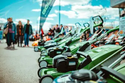 Eine Reihe von grünen Rasenmähern steht im Freien auf einem Bürgersteig mit Preisschildern, wie in einer aktuellen Pressemitteilung zu sehen ist. Im Hintergrund gehen Menschen spazieren und versammeln sich unter einem blauen Himmel mit Wolken, während der Fokus auf den Rasenmähern bleibt.