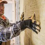 Eine Person, die eine Schutzbrille, eine Mütze und Handschuhe trägt, bringt Dämmmaterial an einer Wand an und drückt es mit beiden Händen fest. Die Isolierung scheint gelb und faserig zu sein.
