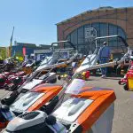 Auf einer Maschinenausstellung sind mehrere orangefarbene Rasenmäher mit Preisschildern im Freien ausgestellt. Eine Person steht in der Nähe, und im Hintergrund ist ein großes Backsteingebäude mit einem Glaseingang zu sehen.