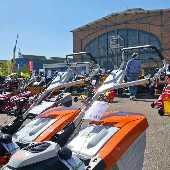 Auf einer Maschinenausstellung sind mehrere orangefarbene Rasenmäher mit Preisschildern im Freien ausgestellt. Eine Person steht in der Nähe, und im Hintergrund ist ein großes Backsteingebäude mit einem Glaseingang zu sehen.