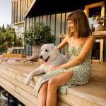 Eine Frau in einem grünen, geblümten Kleid sitzt auf einer hölzernen Veranda, lächelt und streichelt einen großen weißen Hund. Auf der Veranda stehen Topfpflanzen und ein Tablett mit Blumen, im Hintergrund sind Grünpflanzen und ein modernes Haus zu sehen.