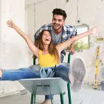 Eine lächelnde Frau sitzt mit ausgestreckten Armen in einer Schubkarre, während ein Mann hinter ihr steht und die Karre schiebt. Sie wirken glücklich und verspielt in einem Raum, der gerade renoviert wird.