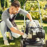 Ein Mann mit Schürze und Handschuhen kniet auf dem Rasen, während er in einem sonnigen Garten einen Rasenmäher wartet.