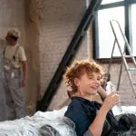 Eine Frau mit gelocktem rotem Haar entspannt sich auf einem mit Plastik überzogenen Sofa, lächelt und hält eine Wasserflasche. Im Hintergrund streicht eine Person in einem sonnenbeschienenen Raum mit einer Leiter in der Nähe eine Ziegelwand.