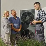 Ein Techniker in einem Overall spricht mit einem lächelnden Paar im Freien, neben einer Wärmepumpeneinheit, die an einer Gebäudewand montiert ist, mit lila Blumen im Vordergrund.