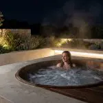 Eine Frau entspannt sich nachts in einem Holzbottich im Freien, umgeben von Steinmauern und beleuchteten Pflanzen, während Dampf aus dem Wasser aufsteigt.