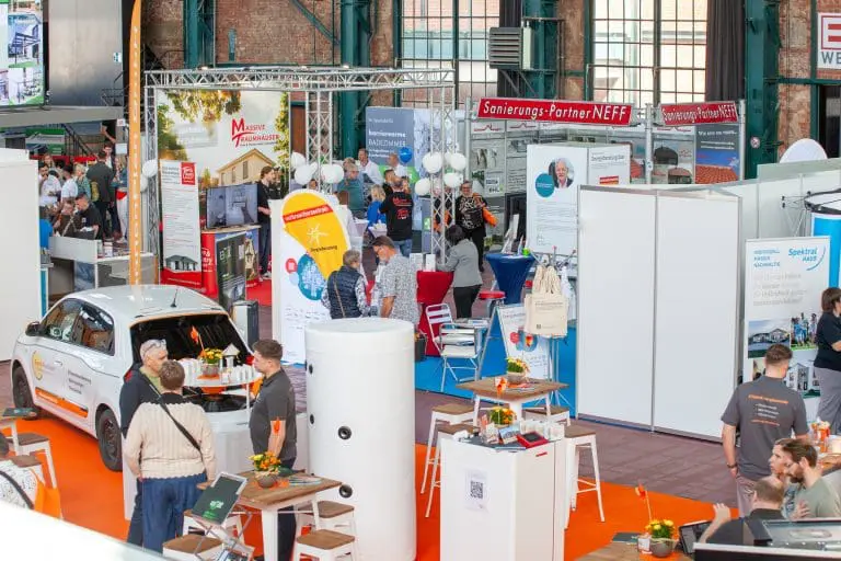 Eine belebte überdachte Messe mit Menschen, die in der Nähe von Ständen, Auslagen und einem weißen Auto herumlaufen und sich unterhalten. Bunte Banner, Informationsplakate und Tische mit Broschüren füllen die Szene. Die Umgebung wirkt lebendig und professionell.