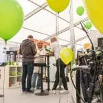Drei Personen stehen um einen hohen Tisch herum und unterhalten sich in einem hellen Messestand. Im Hintergrund sind grüne Luftballons und Ausstellungsregale mit verschiedenen Artikeln zu sehen.