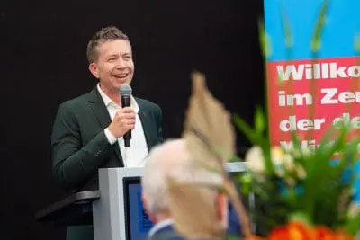 Ein Mann in einem grünen Anzug spricht lächelnd in ein Mikrofon an einem Podium. Im Vordergrund sind verschwommene Blumen zu sehen, und hinter ihm ist ein buntes Schild mit deutschem Text zu sehen.