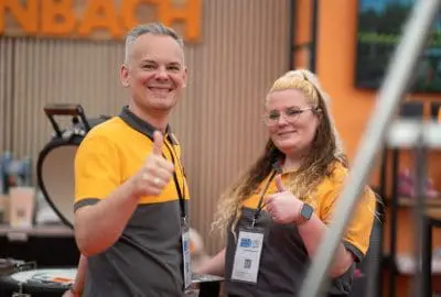 Zwei Personen in passenden orange-grauen Uniformen lächeln und zeigen den Daumen nach oben in die Kamera bei einer Indoor-Veranstaltung. Beide tragen Namensschilder und stehen vor einem unscharfen Hintergrund mit orangefarbener Beschilderung.