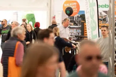Eine geschäftige Veranstaltung in einer Halle, bei der verschiedene Menschen herumlaufen und sich unterhalten. Im Hintergrund sind Stände und Werbebanner zu sehen, und ein Mann in einem karierten Hemd steht hinter einem Tresen mit Broschüren.