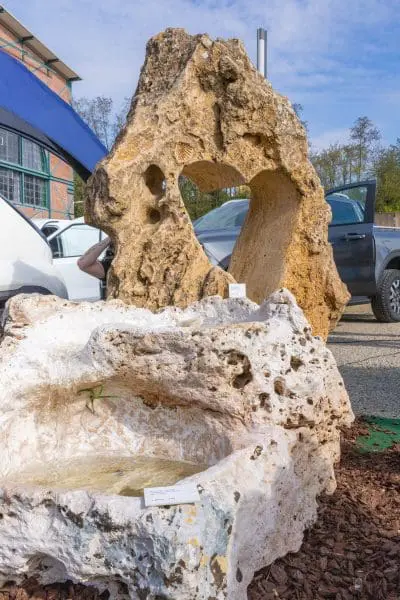 Große dekorative Felsen im Freien; der Felsen im Hintergrund hat ein herzförmiges Loch, während der Felsen im Vordergrund weiß ist und eine flache Schale hat. Im Hintergrund sind Autos und ein Gebäude zu sehen.