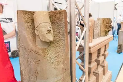 Eine Steinskulptur mit einem geschnitzten menschlichen Gesicht und einem Hut steht aufrecht neben anderen geschnitzten Steinfiguren in einer Innenausstellung mit blauem Boden und Metallstützen.