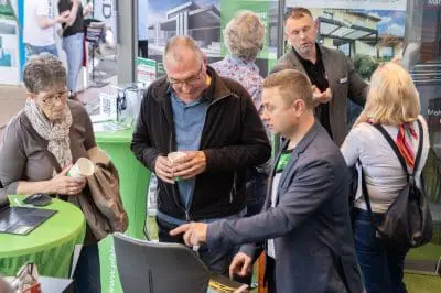 Eine Gruppe von Menschen steht bei einer Veranstaltung in einem Gebäude um einen grünen Tisch herum und unterhält sich. Ein Mann im Anzug zeigt auf einen Bildschirm, während andere zuhören und Tassen halten. Im Hintergrund sind Bilder von modernen Häusern zu sehen.