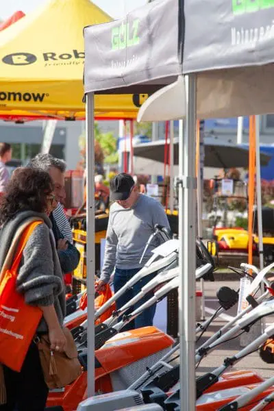 Menschen begutachten auf einer Messe oder Ausstellung Motorgeräte für den Außenbereich. Es sind orangefarbene und graue Maschinen ausgestellt, und im Hintergrund sind Markenzelte und Banner zu sehen. Die Szene wirkt geschäftig und lebendig.