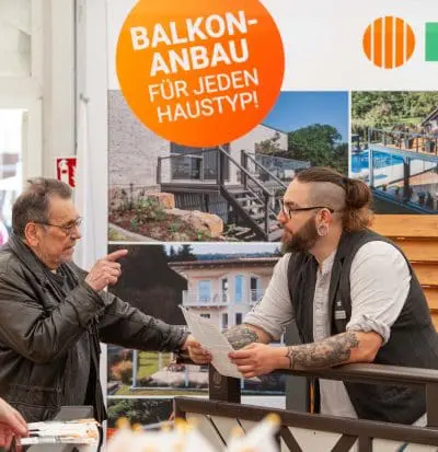 Ein älterer Mann unterhält sich mit einem bärtigen Mitarbeiter, der an einem Stand mit einem Schild "BALKON-ANBAU FÜR JEDEN HAUSTYP!" und Fotos von Balkonen im Hintergrund Papiere hält.