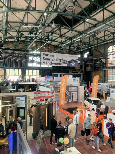 Menschen erkunden verschiedene Stände auf einer überdachten Messe, über denen Banner mit der Aufschrift Forum Saarländisches Handwerk hängen. Der Veranstaltungsort hat hohe Decken, große Fenster und Aussteller, die ihre Produkte und Dienstleistungen vorstellen.