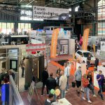 Eine Indoor-Messe mit zahlreichen Ständen, Bannern und Displays. Viele Menschen gehen umher oder sprechen mit den Ausstellern. Auf den Bannern stehen Firmennamen, und auf einem großen Schild steht "Forum Saarländisches Handwerk".