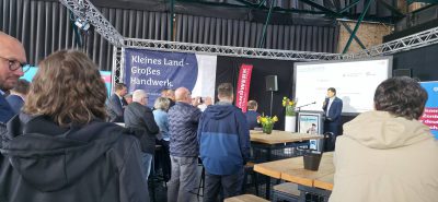 Eine Gruppe von Menschen steht in einer Halle mit hohen Decken und hört einem Redner auf einem Podium zu. Es gibt Banner, Blumen auf Tischen und ein Schild mit der Aufschrift "Kleines Land - Großes Handwerk".