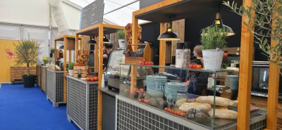 Lebensmittelmarktstand mit Holztheken, Körben mit Brot, frischem Gemüse und Topfpflanzen. In den Vitrinen stehen Baguettes und Schalen. Über der Theke hängen Deckenlampen; hinter den Ständen arbeiten Menschen in einem überdachten Raum mit blauem Boden.