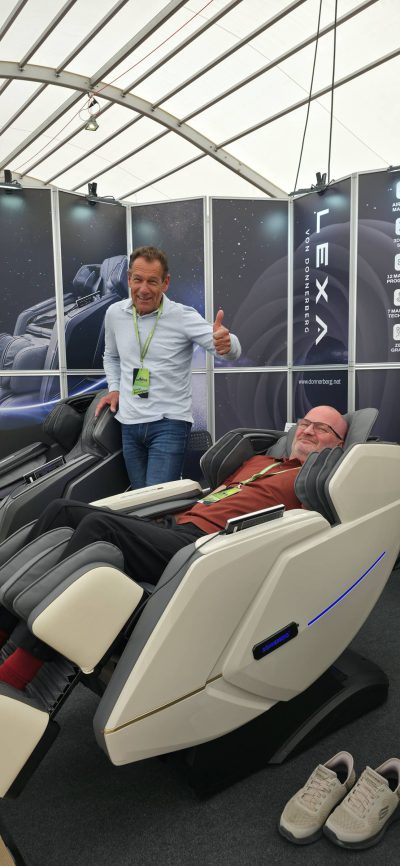 Ein Mann steht lächelnd und mit erhobenem Daumen neben einem anderen Mann, der sich in einem weißen Massagesessel an einem Messestand in einer Halle zurücklehnt.