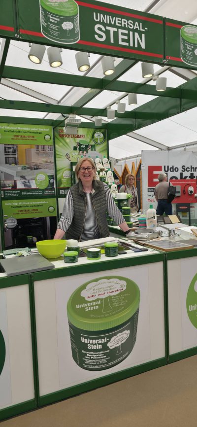 Eine Frau steht hinter einer Theke an einem Stand von Universal-Stein und stellt Reinigungsprodukte aus. Sie lächelt, trägt eine Brille und eine graue Weste. Der Stand ist mit Produktproben, Informationsschildern und grünem Branding ausgestattet.