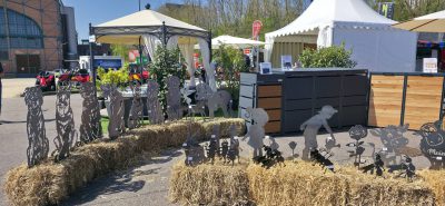 Gartendekorationen aus Metall in Form von Menschen und Tieren werden auf Strohballen auf einem Markt im Freien ausgestellt. Im Hintergrund sind weiße Zelte, Pflanzen und Holzschränke unter einem sonnigen Himmel aufgebaut.