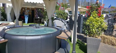 Ein runder Whirlpool mit sprudelndem Wasser steht unter einer Überdachung auf einer Outdoor-Ausstellung. Pflanzen in Töpfen, Terrassenmöbel und Ausstellungsstände sind um den Whirlpool herum zu sehen, während die Besucher im Hintergrund stöbern.