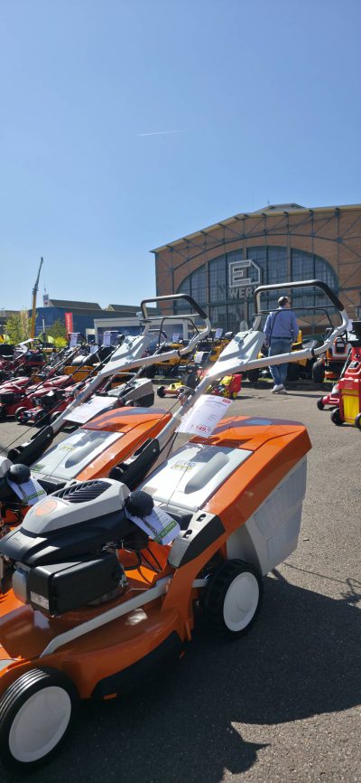 An einem sonnigen Tag sind vor einem großen Gebäude Reihen von bunten Rasenmähern und Gartengeräten ausgestellt, zwischen denen ein paar Leute spazieren gehen.