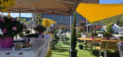 Sitzbereich im Freien mit Tischen und Stühlen unter großen Sonnenschirmen, umgeben von Topfpflanzen und Blumen in bunten Töpfen, auf Kunstrasen, mit einem Gebäude und Bäumen im Hintergrund an einem sonnigen Tag.