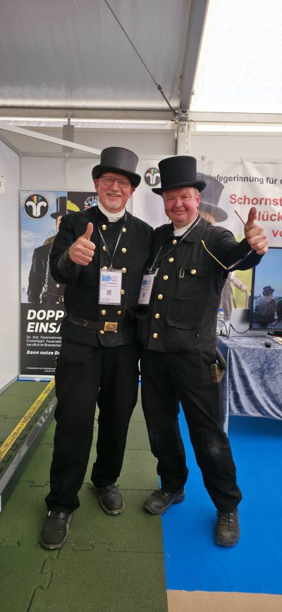 Zwei lächelnde Männer in traditionellen schwarzen Schornsteinfegeruniformen und Zylinderhüten stehen nebeneinander und zeigen die Daumen nach oben in einem Veranstaltungsstand mit Bannern und blauem Bodenbelag.