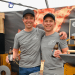 Zwei lächelnde Männer in passenden grauen Hemden und schwarzen Mützen stehen nebeneinander und halten jeweils ein Getränk in der Hand. Sie stehen vor einem Stand mit einer feurigen Kulisse und Grillgeräten und wirken glücklich und entspannt.