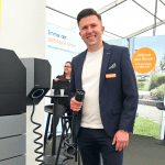 Ein lächelnder Mann in einem blauen Anzug hält einen Ladestecker für Elektroautos an einem Messestand. Hinter ihm sitzt eine Frau an einem Tisch, und es sind Informationsplakate über Energie und Strom zu sehen.