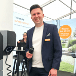 Ein Mann in einem blauen Anzug lächelt, während er einen Ladestecker für Elektrofahrzeuge an einem Stand hält, an dem für erschwingliche und intelligente Energielösungen geworben wird. Im Hintergrund sind Werbeplakate zu sehen.