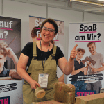 Eine lächelnde Frau in Latzhose und Schutzbrille zeigt an einem Verkaufsstand den Daumen nach oben und hält einen Hammer in der Nähe eines behauenen Steins. Auf deutschen Bannern hinter ihr steht "Spaß am Wir?" und wirbt für die Ausbildung zum Steinmetz.