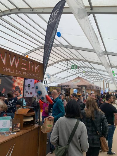 Eine Menschenmenge geht durch eine belebte Markthalle oder Messe mit verschiedenen Verkaufsständen. Eine Person in einem Raketenmaskottchen-Kostüm steht in der Nähe eines Standes, und über den Ständen sind Banner und Displays zu sehen.