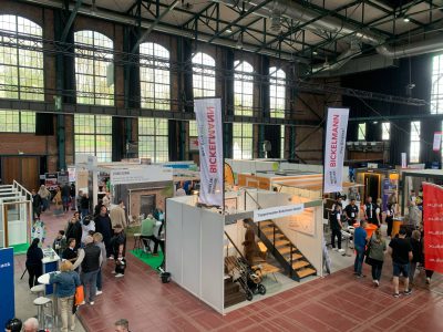 Eine geschäftige Indoor-Messe mit verschiedenen Ständen, Bannern und Displays. In einer geräumigen Industriehalle mit hohen Decken laufen die Menschen umher, interagieren und begutachten die Exponate unter hohen Fenstern.
