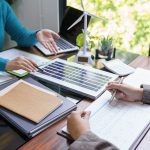 Zwei Personen besprechen Dokumente an einem Schreibtisch mit einem Laptop, einem Modell-Windrad, einem kleinen Solarpanel, einem Taschenrechner und Topfpflanzen, was auf eine Besprechung über erneuerbare Energien hindeutet.