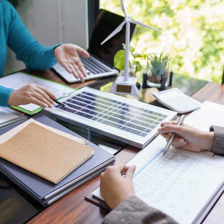 Zwei Personen besprechen Dokumente an einem Schreibtisch mit einem Laptop, einem Modell-Windrad, einem kleinen Solarpanel, einem Taschenrechner und Topfpflanzen, was auf eine Besprechung über erneuerbare Energien hindeutet.