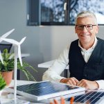 Ein lächelnder Mann mit Brille sitzt an einem Schreibtisch mit einem Solarmodell, einem Windradmodell und einer Topfpflanze in einem hellen Büro mit großen Fenstern im Hintergrund.