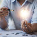 Eine Person in einem hellblauen Hemd hält eine leuchtende Glühbirne in der einen und einen Stift in der anderen Hand, während vor ihr auf dem Schreibtisch Papiere liegen.